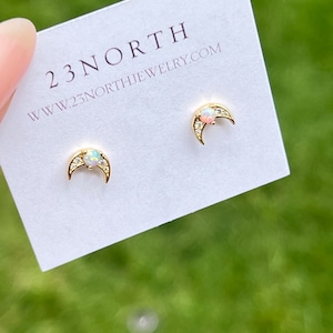 May include: A pair of gold crescent moon earrings with a small opal stone in the center of each moon. The earrings are on a white card with the text "23NORTH www.23northjewelry.com".