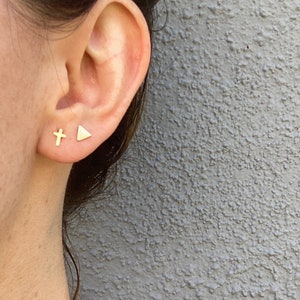 May include: A close-up of a person's ear with two gold stud earrings. One earring is a small cross, and the other is a triangle. The earrings are set against a gray textured background.
