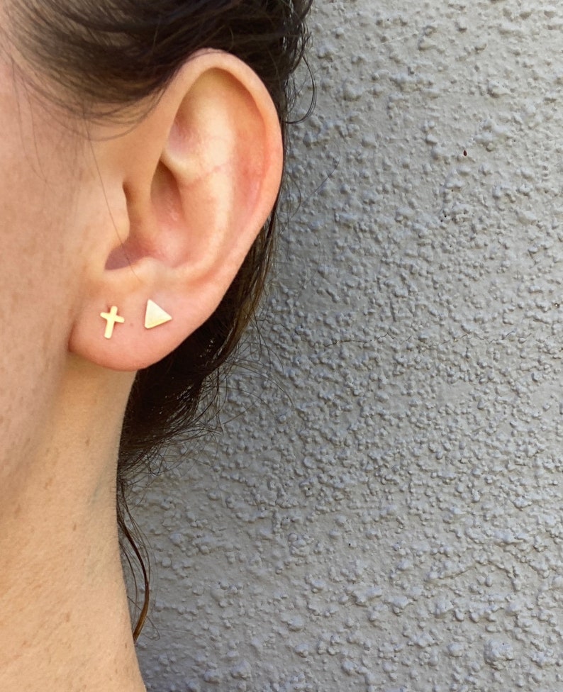 May include: A close-up of a person's ear with two gold stud earrings. One earring is a small cross, and the other is a triangle. The earrings are set against a gray textured background.