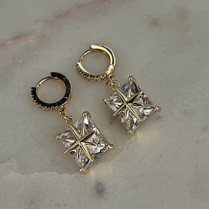 Puede incluir: Un par de pendientes de aro dorados. Cada pendiente presenta una gema cuadrada y transparente con un diseño de estrella dorada en el centro. Los aros también están adornados con pequeñas gemas transparentes.