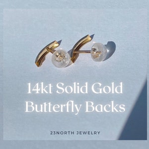 May include: Two 14kt solid gold butterfly earring backs with clear plastic stoppers. The gold pieces have a curved design. The text "14kt Solid Gold Butterfly Backs" is displayed on a light blue background. The brand name "23NORTH JEWELRY" is also visible.