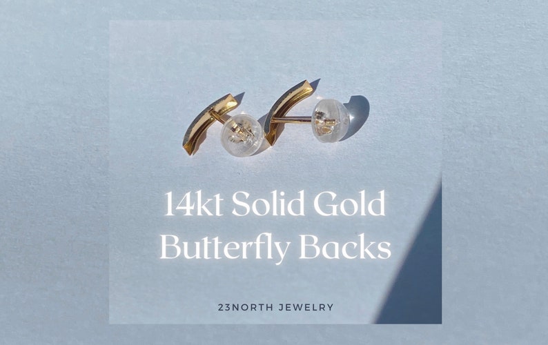 May include: Two 14kt solid gold butterfly earring backs with clear plastic stoppers. The gold pieces have a curved design. The text "14kt Solid Gold Butterfly Backs" is displayed on a light blue background. The brand name "23NORTH JEWELRY" is also visible.