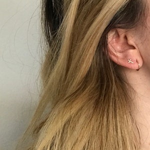 May include: Close-up of a person's ear adorned with two small gold earrings. One is a cross shape, and the other is a small hoop. The person has long, wavy blonde hair with darker roots.