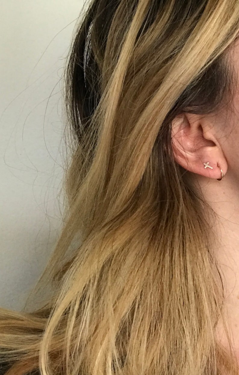 May include: Close-up of a person's ear adorned with two small gold earrings. One is a cross shape, and the other is a small hoop. The person has long, wavy blonde hair with darker roots.