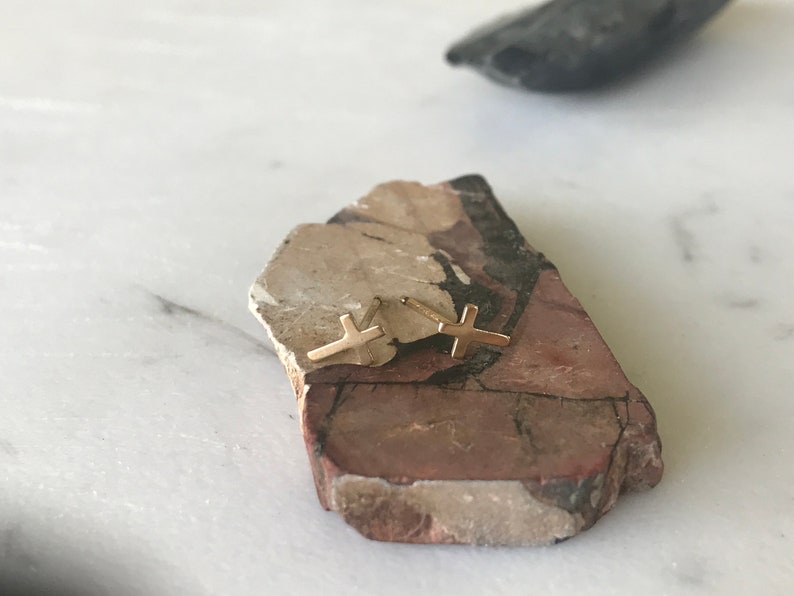 May include: A pair of gold-colored cross stud earrings. The earrings are displayed on a multi-colored, textured stone, set against a white marble surface. The cross design is simple and elegant.