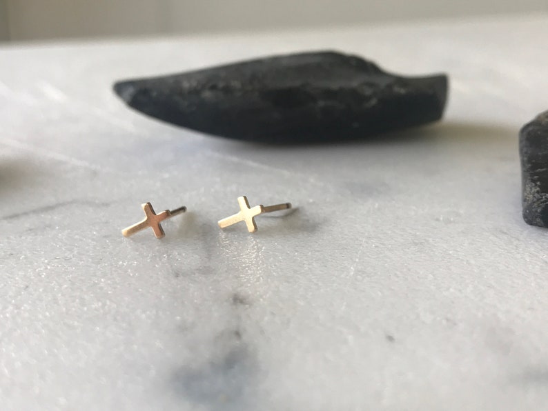 May include: A pair of small, gold-colored cross stud earrings. The earrings are displayed on a white and gray marble surface, with a dark, irregularly shaped object in the background. The earrings are a simple, minimalist design.