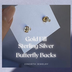 May include: Close-up of two gold-filled butterfly earring backs on a white card. The text on the card reads "Gold Fill Sterling Silver Butterfly Backs" and "23NORTH JEWELRY".