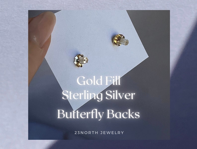 May include: Close-up of two gold-filled butterfly earring backs on a white card. The text on the card reads "Gold Fill Sterling Silver Butterfly Backs" and "23NORTH JEWELRY".