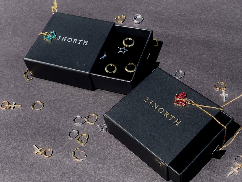 May include: Black jewelry boxes with the text "23 NORTH" and various earrings and a necklace. The earrings include hoops, star-shaped, and cross designs. The necklace features a red pendant. The jewelry is gold and silver.