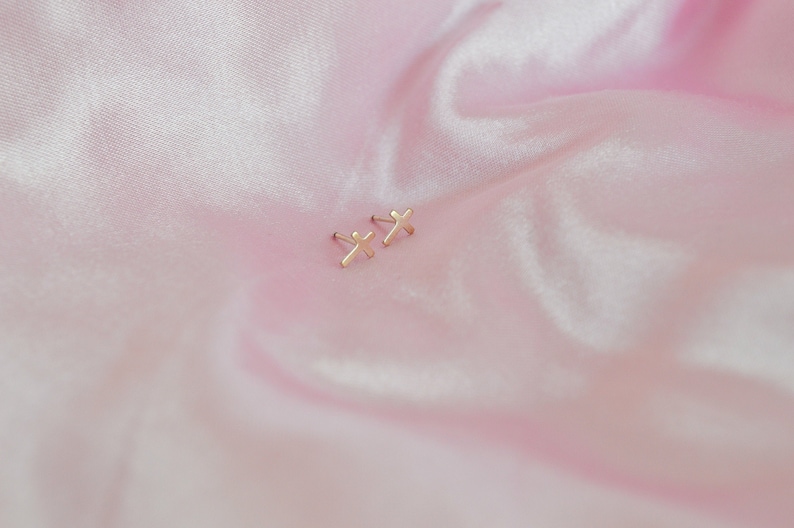 May include: A pair of small, gold-colored cross stud earrings. The earrings are displayed on a pink, satin-like fabric. The crosses are simple in design and appear to be made of metal.