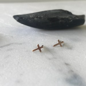 May include: A pair of small, gold-colored cross stud earrings. The earrings are displayed on a white marble surface, with a dark, oblong stone in the background. The crosses are simple in design.