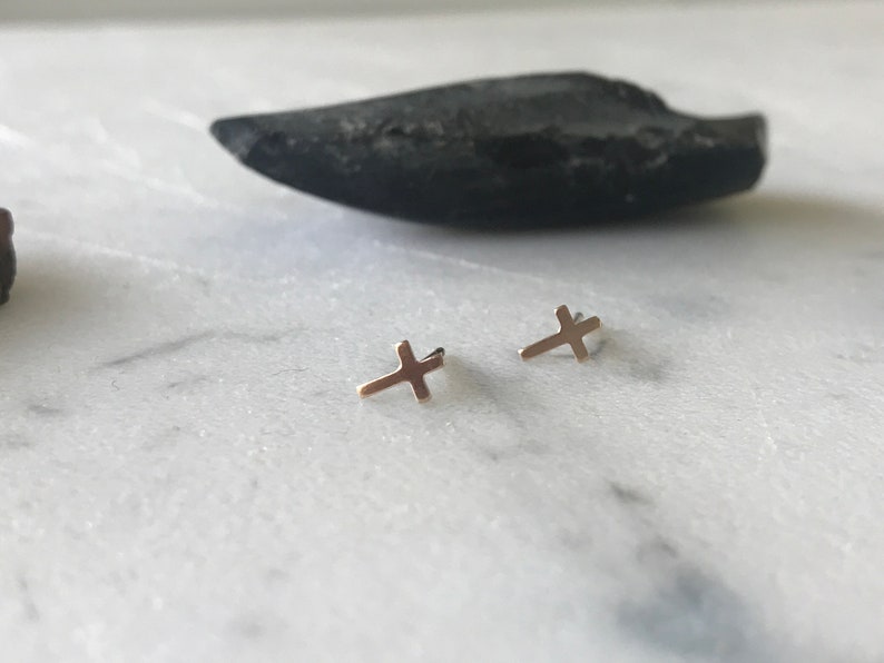 May include: A pair of small, gold-colored cross stud earrings. The earrings are displayed on a white marble surface, with a dark, oblong stone in the background. The crosses are simple in design.