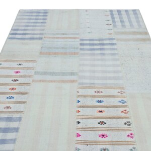 May include: A rectangular, patchwork rug with a cream-colored base. The rug features various rectangular sections with stripes in blue, tan, and gray, and sections with floral patterns in pink, blue, and orange. The rug is made of a woven material.