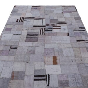 Puede incluir: Una alfombra grande y rectangular de patchwork con una paleta de colores neutros. La alfombra está compuesta por numerosos parches rectangulares y cuadrados en tonos grises, beige y marrón. Algunos parches presentan rayas marrones oscuras. Adecuada para salón o dormitorio.
