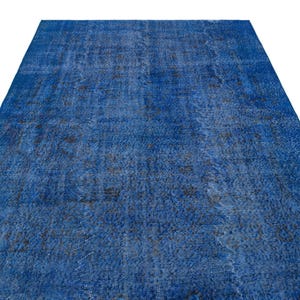 Puede incluir: Una alfombra azul rectangular grande con una apariencia desgastada y vintage. La alfombra tiene un patrón descolorido y moteado con áreas más oscuras, creando una apariencia texturizada. La alfombra se muestra sobre un fondo blanco.