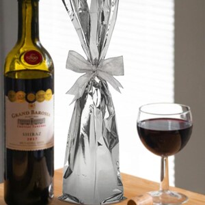 May include: A silver foil gift bag with a white ribbon tied around it. The bag is sitting on a wooden surface next to a bottle of wine, a glass of wine, and a cork.