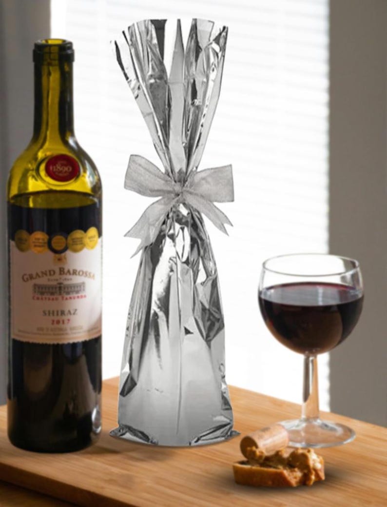 May include: A silver foil gift bag with a white ribbon tied around it. The bag is sitting on a wooden surface next to a bottle of wine, a glass of wine, and a cork.
