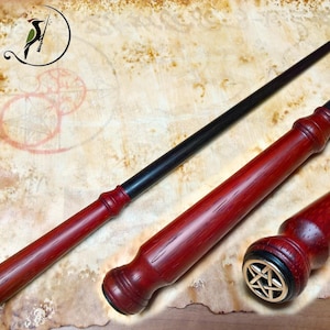 Puede incluir: Una varita de madera hecha a mano con un mango rojo oscuro y una punta negra. El mango presenta un diseño detallado y un emblema dorado. La varita se muestra sobre una superficie similar a un pergamino.