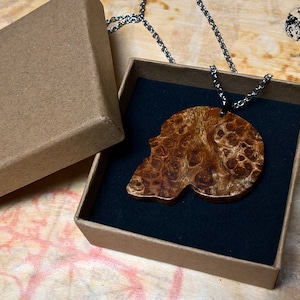 Può includere: Una collana con un ciondolo a forma di teschio in legno in una scatola marrone con fodera in velluto nero. Il ciondolo è in legno con un motivo a radica marrone e bianco. La collana è realizzata con una catena d'argento.