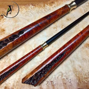 Può includere: Due bacchette artigianali con manici in legno radica lucidato caratterizzati da profonde crepe naturali scure e strutturate. Ogni manico è collegato a un'asta nera sottile e liscia tramite un collare metallico color oro. Logo di uccello visibile.