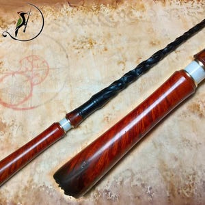 Op de afbeelding: Een lange, slanke zwarte toverstaf met een gedraaide, spiraalvormige schacht en een gladde, gepolijste roodbruine houten handgreep. De greep heeft een cilindrische vorm met een witte band en een metalen ring, overgaand in de donkere punt.