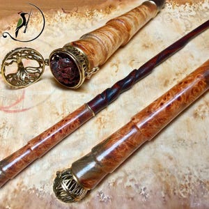 May include: Two wooden wands with polished, swirling grain patterns and metallic gold-toned filigree caps. One wand features a detachable cap with a circular tree-of-life design, revealing a dark, textured interior compartment.