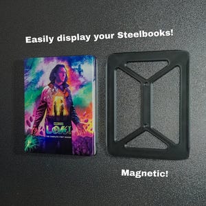 May include: A black metal magnetic stand for displaying Steelbooks. The stand has a rectangular shape with a grid pattern. The text "Magnetic!" is written below the stand. The stand is shown next to a Steelbook case featuring the Marvel series "Loki" with the text "The Complete First Season" on the front.