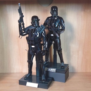 May include: Two black Death Trooper action figures from Star Wars, standing on black display bases. The figures are highly detailed, with helmets, armour, and weapons. One figure holds a blaster rifle, and the other has a blaster rifle at their side.
