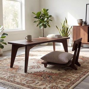 Peut inclure: Une table basse rectangulaire en bois à profil bas avec des pieds inclinés et une chaise de sol assortie dotée d'un dossier et d'un coussin gris clair. La table porte une tasse en céramique sur un dessous de verre, posée sur un tapis.
