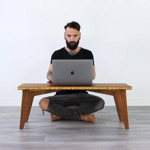 May include: A person sits cross-legged on the floor in front of a wooden table, using a laptop computer. The table has a natural wood finish and a simple design.