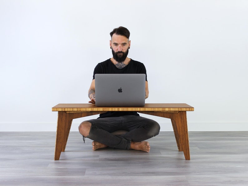 May include: A person sits cross-legged on the floor in front of a wooden table, using a laptop computer. The table has a natural wood finish and a simple design.