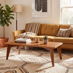 May include: A rectangular wooden coffee table with angled legs sits on a cream-colored rug with a geometric pattern. A mustard yellow sofa with a beige throw blanket and two patterned throw pillows is positioned behind the coffee table.
