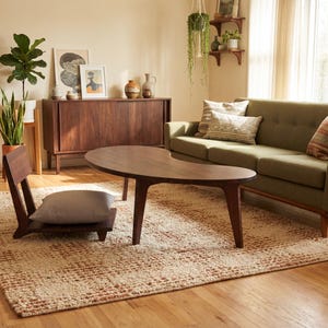 May include: A low-profile wooden floor chair with a light gray cushion, positioned on a textured cream and brown area rug. The chair features a simple backrest and a flat seat, placed near a kidney-shaped wooden coffee table in a living room.
