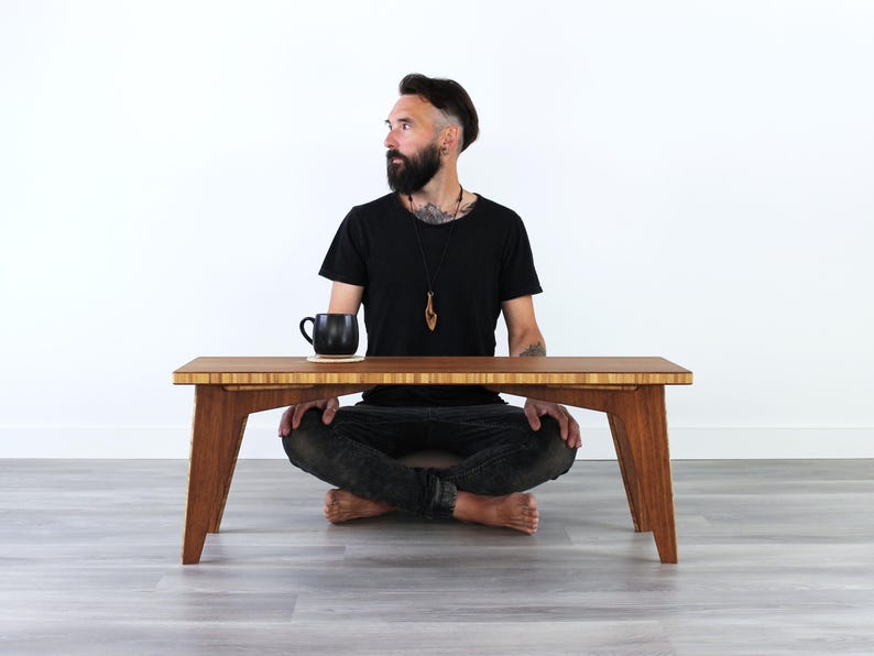 May include: A person sits cross-legged on the floor in front of a wooden table. The table has a light brown finish and a rectangular shape. A black mug sits on a coaster on the table.