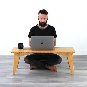 May include: A person sits cross-legged on the floor in front of a light wood table. They are using a silver laptop computer. A black mug sits on the table.