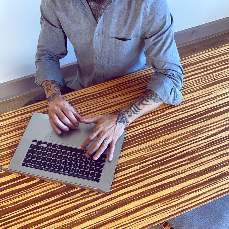 May include: A person with tattoos on their arms is typing on a silver laptop computer with a black keyboard. The laptop is on a wooden table with a striped pattern.