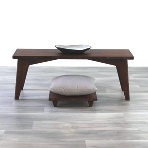 May include: A dark brown wooden bench with a light gray cushion on a small wooden platform. The bench has a simple design with straight legs and a rectangular top. A white ceramic bowl sits on top of the bench.