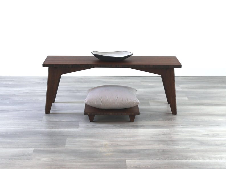 May include: A dark brown wooden bench with a light gray cushion on a small wooden platform. The bench has a simple design with straight legs and a rectangular top. A white ceramic bowl sits on top of the bench.
