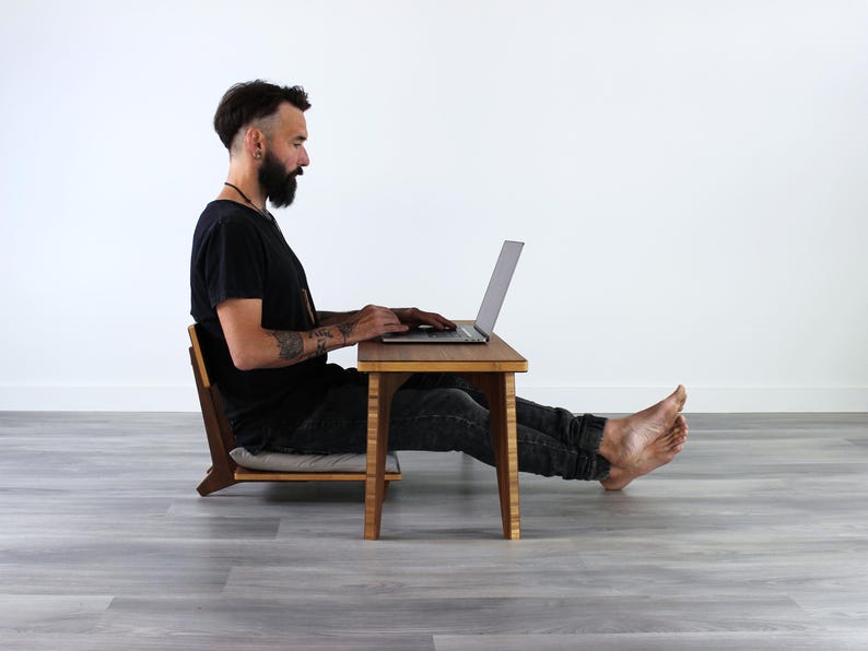 May include: A person sits on a wooden chair with a cushion and uses a laptop on a wooden table. The table and chair are both made of natural wood and have a simple, minimalist design.