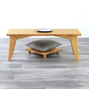 May include: A light wood coffee table with a gray cushion on a small wooden stand. The table has a simple design with straight legs and a rectangular top.