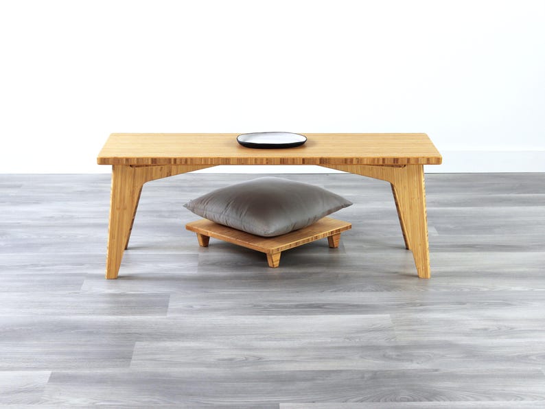 May include: A light wood coffee table with a gray cushion on a small wooden stand. The table has a simple design with straight legs and a rectangular top.