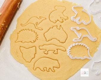 May include: A close-up of a sheet of cookie dough with various animal-shaped cookie cutters pressed into it. The cutters include a koala, a kangaroo, a platypus, a hedgehog, and a bear. The dough is a light yellow colour and the cutters are white.