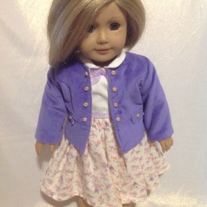 May include: A doll wearing a purple velvet jacket with white buttons, a white collared shirt with a lavender bow, and a white and lavender floral print skirt. The doll has blonde hair and brown eyes.