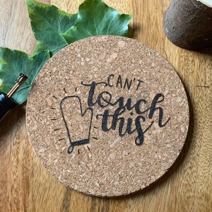 May include: A round cork coaster featuring a wood-burned design of an oven mitt and the text "CAN'T touch this" in black lettering. The coaster is positioned next to a wood-burning tool and green leaves, showcasing a natural, textured surface.