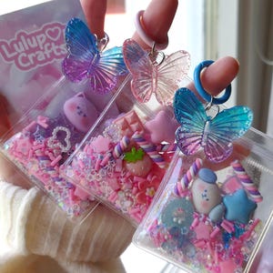 May include: Three clear, rectangular keychains filled with colorful sprinkles and small charms. Each keychain has a butterfly charm and a ring. The keychains are held in front of a window. The brand name "Lulup Crafts" is visible.