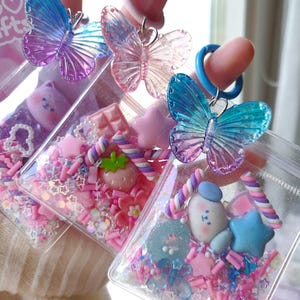 May include: Three clear, square-shaped charms filled with pink and white sprinkles, small candies, and decorative elements. Each charm has a different design, including a strawberry, a bear, and a star. Each charm is topped with a translucent butterfly.