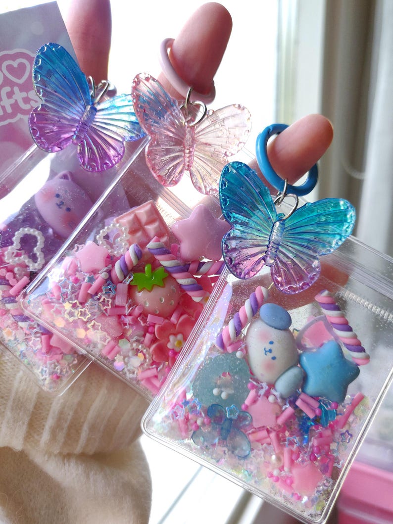 May include: Three clear, square-shaped charms filled with pink and white sprinkles, small candies, and decorative elements. Each charm has a different design, including a strawberry, a bear, and a star. Each charm is topped with a translucent butterfly.