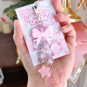 May include: A pink keychain with a bow, stars, and a flower charm. The keychain is attached to a card with the text "LuluPop Crafts" and social media handles. The design features a silver clasp and various pink beads.
