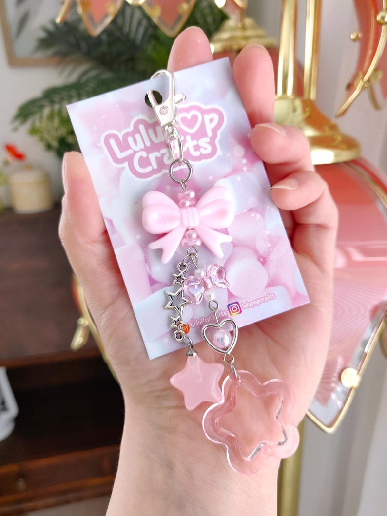 May include: A pink keychain with a bow, stars, and a flower charm. The keychain is attached to a card with the text "LuluPop Crafts" and social media handles. The design features a silver clasp and various pink beads.
