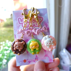 May include: Three adorable donut-shaped keyrings with a bear, chick, and bunny face. Each keyring has a pink bow and a gold chain with a lobster clasp. The keyrings are displayed on a pink and white card with the text "LuluPopCrafts" and a social media icon.